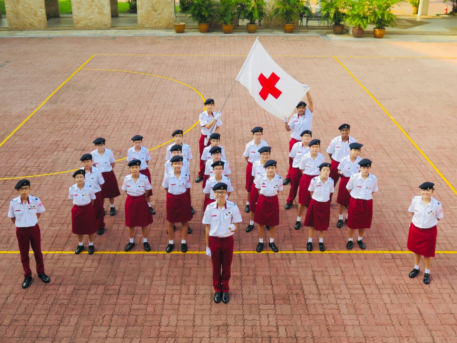 Students in white and red uniforms with black berets stand in formation on a courtyard, one holding a Red Cross flag.